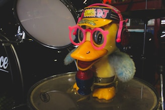 A cartoonish duck figure with bright yellow body, wearing oversized pink glasses and blue earmuffs with a colorful hat labeled 'Duck DJ'. It stands on top of a drum in a music-themed setting with other drum parts visible in the background.