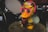 A cartoonish duck figure with bright yellow body, wearing oversized pink glasses and blue earmuffs with a colorful hat labeled 'Duck DJ'. It stands on top of a drum in a music-themed setting with other drum parts visible in the background.