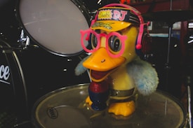 A cartoonish duck figure with bright yellow body, wearing oversized pink glasses and blue earmuffs with a colorful hat labeled 'Duck DJ'. It stands on top of a drum in a music-themed setting with other drum parts visible in the background.