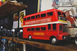 A detailed model of a red London double-decker bus is placed on a wooden shelf. The bus is labeled 'London City Sightseeing' and has the number 8 route, listing notable destinations on its side. Surrounding the bus are vintage cameras and a variety of eclectic items, including colorful art and posters on the wall behind.