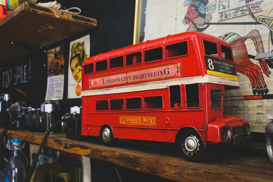 A detailed model of a red London double-decker bus is placed on a wooden shelf. The bus is labeled 'London City Sightseeing' and has the number 8 route, listing notable destinations on its side. Surrounding the bus are vintage cameras and a variety of eclectic items, including colorful art and posters on the wall behind.