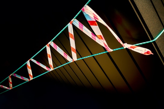 Brightly colored caution tape with red and white stripes is tied along a green rope against a dark background. The tape casts sharp shadows, creating a striking contrast and dynamic pattern.