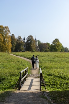A couple walks hand-in-hand along a narrow path in a grassy field, leading towards a picturesque church with distinct spires in the distance. The scene is framed by lush green trees on either side and a simple wooden bridge in the foreground. The clear sky and the warm colors of the foliage suggest it's a serene day, likely in early autumn.