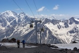 Two hikers are walking on a snow-covered mountain path with trekking poles. In the distance, majestic snow-capped mountains rise against a clear blue sky. A ski lift with cable cars traverses the scene, adding a sense of adventure and exploration.