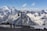 Two hikers are walking on a snow-covered mountain path with trekking poles. In the distance, majestic snow-capped mountains rise against a clear blue sky. A ski lift with cable cars traverses the scene, adding a sense of adventure and exploration.