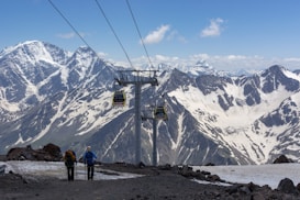 Two hikers are walking on a snow-covered mountain path with trekking poles. In the distance, majestic snow-capped mountains rise against a clear blue sky. A ski lift with cable cars traverses the scene, adding a sense of adventure and exploration.