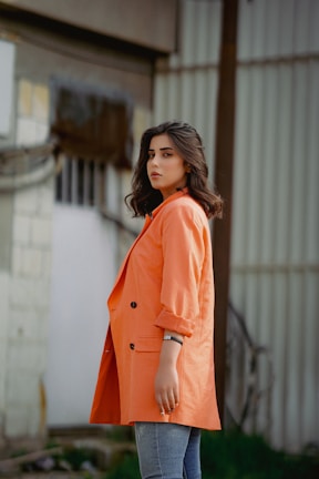 A woman stands in a side pose, wearing an oversized orange coat and denim pants. Her hair is shoulder-length and styled wavy. Behind her is a background of industrial elements, including metal and concrete surfaces.