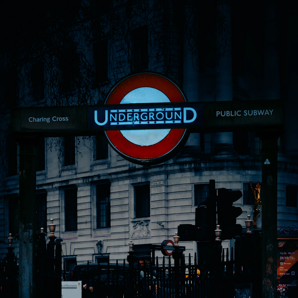 The image shows a street scene at night, with a large sign for the Underground prominently displayed on a building. The sign is illuminated by blue lights and features the words "Underground" written in white letters against a red background. Below the sign, there are two traffic lights visible - one on the left side of the frame and another further to the right. In front of the building, there's a car parked along with several people walking around or standing nearby. The scene is set against a dark sky, creating an atmosphere of mystery and intrigue.