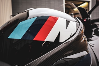 A close-up of a sleek black surface featuring the iconic M logo in blue, dark blue, and red stripes. The surface appears to be part of a vehicle, possibly a sports car or motorcycle, with reflections visible in the glossy finish.