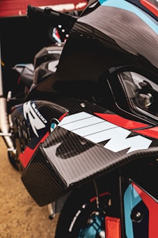 Close-up of a motorcycle with a focus on the intricate details of its carbon fiber components and distinctive M branding. The bike features a combination of sleek, aerodynamic lines and vibrant colors, including red, blue, and black.
