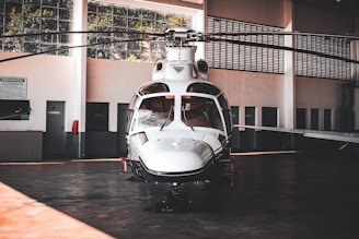 A sleek white helicopter is parked inside a spacious hangar. The hangar features large windows that allow natural light to filter through, illuminating the aircraft. The helicopter's design is modern with a shiny exterior surface, and its rotors are still. Surrounding the helicopter are the hangar's walls, doors, and some signs, giving an industrial and functional atmosphere.