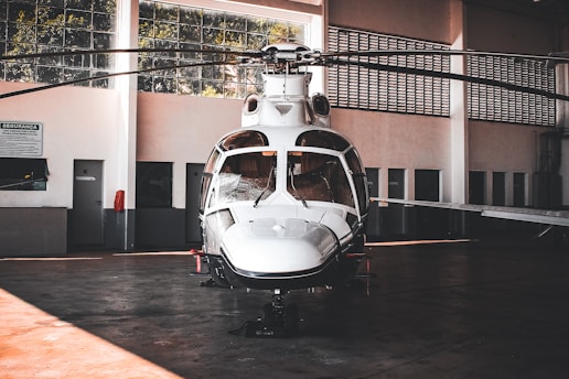 A sleek white helicopter is parked inside a spacious hangar. The hangar features large windows that allow natural light to filter through, illuminating the aircraft. The helicopter's design is modern with a shiny exterior surface, and its rotors are still. Surrounding the helicopter are the hangar's walls, doors, and some signs, giving an industrial and functional atmosphere.