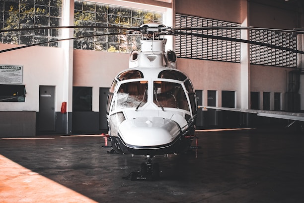 A sleek white helicopter is parked inside a spacious hangar. The hangar features large windows that allow natural light to filter through, illuminating the aircraft. The helicopter's design is modern with a shiny exterior surface, and its rotors are still. Surrounding the helicopter are the hangar's walls, doors, and some signs, giving an industrial and functional atmosphere.