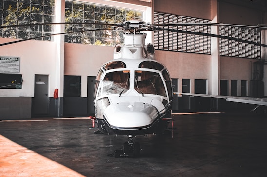 A sleek white helicopter is parked inside a spacious hangar. The hangar features large windows that allow natural light to filter through, illuminating the aircraft. The helicopter's design is modern with a shiny exterior surface, and its rotors are still. Surrounding the helicopter are the hangar's walls, doors, and some signs, giving an industrial and functional atmosphere.