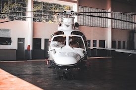A sleek white helicopter is parked inside a spacious hangar. The hangar features large windows that allow natural light to filter through, illuminating the aircraft. The helicopter's design is modern with a shiny exterior surface, and its rotors are still. Surrounding the helicopter are the hangar's walls, doors, and some signs, giving an industrial and functional atmosphere.