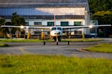 A small aircraft is situated on a runway in front of an industrial building, with greenery partially surrounding the area. The scene is captured during daylight with sunlight reflecting off the plane's wings.