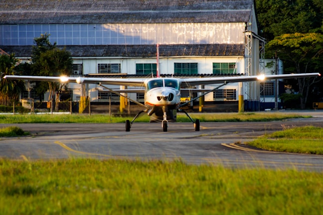 A small aircraft is situated on a runway in front of an industrial building, with greenery partially surrounding the area. The scene is captured during daylight with sunlight reflecting off the plane's wings.
