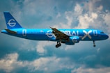 A bright blue airplane with the words 'GLOBAL X' on its side is flying against a partly cloudy sky. The plane appears to be a commercial passenger jet with its landing gear extended.