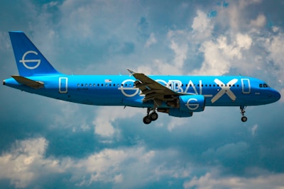 A bright blue airplane with the words 'GLOBAL X' on its side is flying against a partly cloudy sky. The plane appears to be a commercial passenger jet with its landing gear extended.