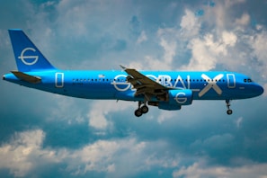 A bright blue airplane with the words 'GLOBAL X' on its side is flying against a partly cloudy sky. The plane appears to be a commercial passenger jet with its landing gear extended.