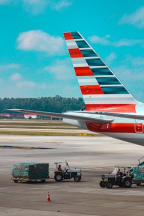 A close-up view of an airplane's tail fin with distinctive red, white, and blue stripes. Below, several small vehicles are towing carts loaded with green-covered cargo. The scene is set at an airport runway with a clear blue sky overhead.