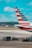 A close-up view of an airplane's tail fin with distinctive red, white, and blue stripes. Below, several small vehicles are towing carts loaded with green-covered cargo. The scene is set at an airport runway with a clear blue sky overhead.