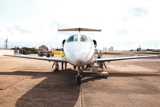 A private jet is parked on an airport tarmac with a refueling truck nearby. Several people, likely ground crew, are seen around the aircraft, working on or inspecting it. The background includes a clear sky and some distant city buildings.