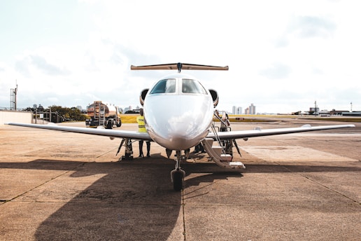 A private jet is parked on an airport tarmac with a refueling truck nearby. Several people, likely ground crew, are seen around the aircraft, working on or inspecting it. The background includes a clear sky and some distant city buildings.