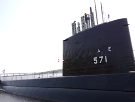 A large black submarine is docked by the water. Numbers and letters, '571' and 'AE', are prominently displayed on the side. The background shows a bridge and a distant shoreline. There are flags hoisted on the submarine, including an American flag.