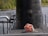 A large, dark metallic structure, possibly the conning tower of a submarine, is situated adjacent to a body of water. It is surrounded by railings and has a coiled rope with orange flotation devices placed on the deck. The background features greenery and a rocky shoreline.