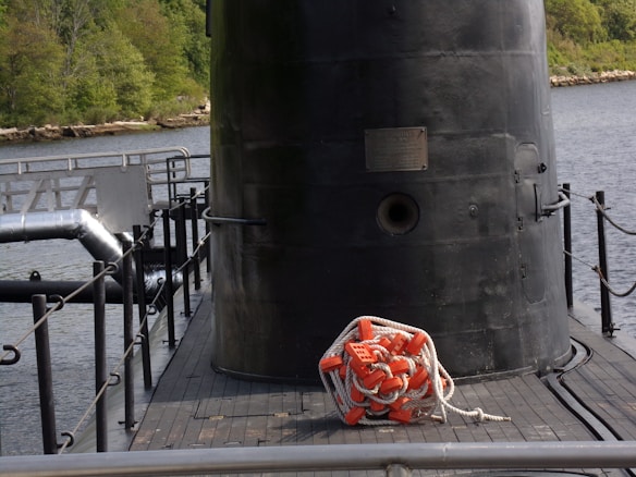 A large, dark metallic structure, possibly the conning tower of a submarine, is situated adjacent to a body of water. It is surrounded by railings and has a coiled rope with orange flotation devices placed on the deck. The background features greenery and a rocky shoreline.