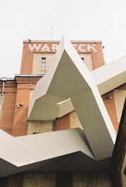 A modern architectural structure with angular lines is juxtaposed against a classic brick building adorned with the word 'WARECK' on its facade. The geometric design of the structure contrasts with the traditional architecture.