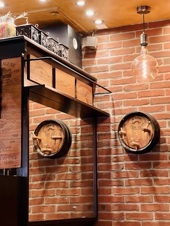 A cozy setting features a brick wall with two wooden barrels mounted on it. Wine crate panels are incorporated into a shelf above a reflective surface. An ornate lamp hangs from the ceiling, casting warm light over the scene. The decor evokes a rustic, vintage atmosphere.