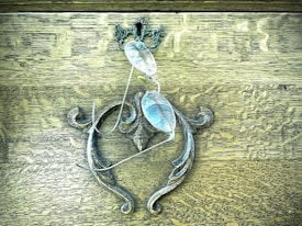 A pair of eyeglasses is hanging on an ornate metal handle attached to a wooden surface. The handle features intricate scrollwork and decorative details, contrasting with the wood's textured grain.