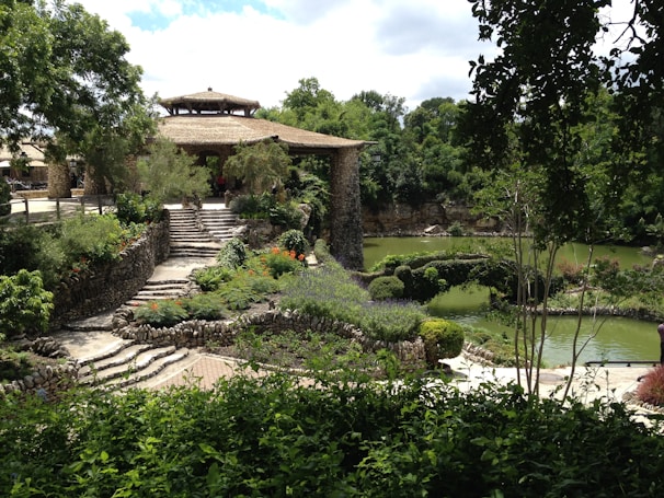 A lush garden scene with carefully landscaped paths and greenery surrounding a stone structure with a thatched roof. The area is abundant with vibrant plants and flowers, bordered by a serene pond reflecting the green foliage. The stone steps and rustic architecture evoke a tranquil and harmonious atmosphere.