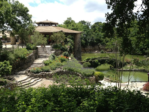 A lush garden scene with carefully landscaped paths and greenery surrounding a stone structure with a thatched roof. The area is abundant with vibrant plants and flowers, bordered by a serene pond reflecting the green foliage. The stone steps and rustic architecture evoke a tranquil and harmonious atmosphere.