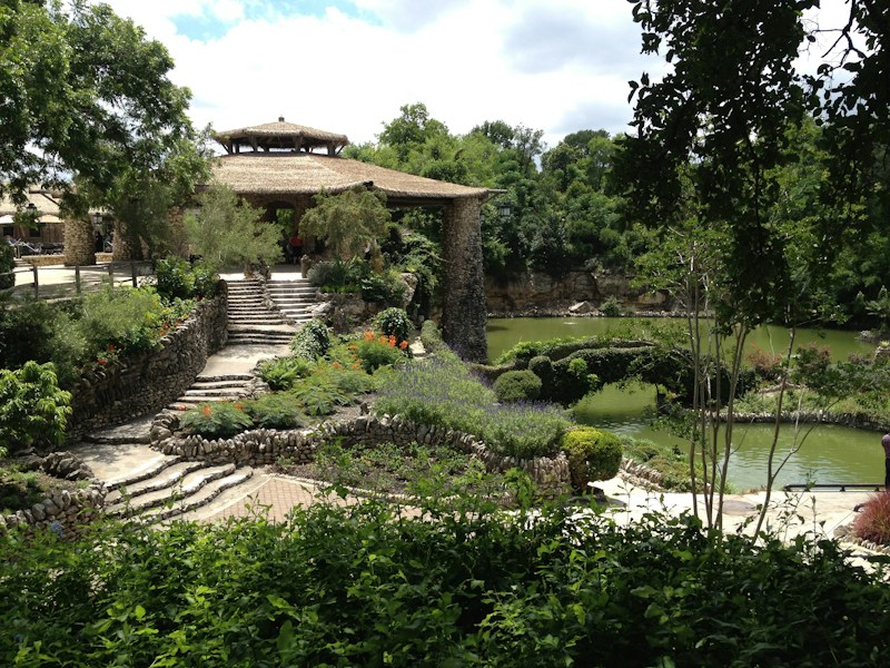 A lush garden scene with carefully landscaped paths and greenery surrounding a stone structure with a thatched roof. The area is abundant with vibrant plants and flowers, bordered by a serene pond reflecting the green foliage. The stone steps and rustic architecture evoke a tranquil and harmonious atmosphere.