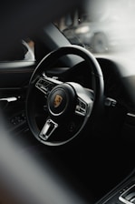 A luxurious car interior featuring a sleek, black leather steering wheel with the Porsche logo prominently displayed in the center. The background is slightly blurred, with the interior reflecting a high-end design.