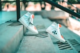 A pair of white high-top sneakers with black arrow designs and bright orange accents is positioned on outdoor concrete steps. The sneakers are styled with black laces and appear to be balancing on the steps, creating a dynamic and modern aesthetic. The background is softly blurred, highlighting the urban setting.