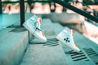 A pair of white high-top sneakers with black arrow designs and bright orange accents is positioned on outdoor concrete steps. The sneakers are styled with black laces and appear to be balancing on the steps, creating a dynamic and modern aesthetic. The background is softly blurred, highlighting the urban setting.