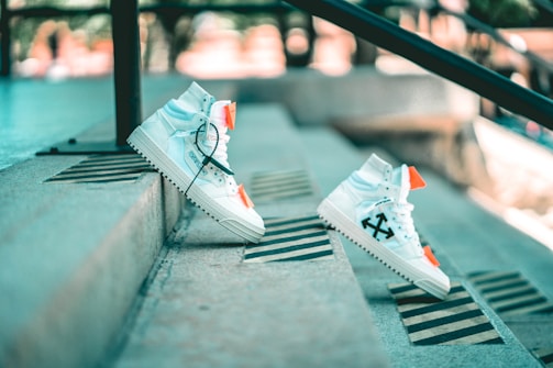 A pair of white high-top sneakers with black arrow designs and bright orange accents is positioned on outdoor concrete steps. The sneakers are styled with black laces and appear to be balancing on the steps, creating a dynamic and modern aesthetic. The background is softly blurred, highlighting the urban setting.