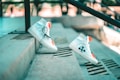 A pair of white high-top sneakers with black arrow designs and bright orange accents is positioned on outdoor concrete steps. The sneakers are styled with black laces and appear to be balancing on the steps, creating a dynamic and modern aesthetic. The background is softly blurred, highlighting the urban setting.