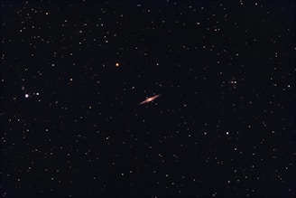 A dark night sky filled with numerous small stars scattered throughout. In the center, a long, thin celestial object, likely a galaxy, stands out with a brighter, elongated appearance.