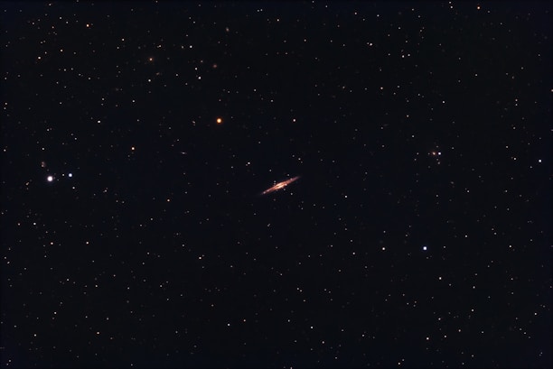 A dark night sky filled with numerous small stars scattered throughout. In the center, a long, thin celestial object, likely a galaxy, stands out with a brighter, elongated appearance.