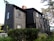 A dark-colored wooden house with a traditional design, featuring multiple windows and a red brick chimney. The surrounding area includes lush greenery, trees, and a glimpse of a body of water. The house has a classic, multi-story architecture.