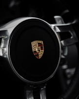 The image features a close-up of a steering wheel with a prominent car emblem at its center. The emblem is a combination of gold and red with detailed iconography, surrounded by polished metallic spokes.