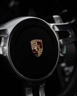 The image features a close-up of a steering wheel with a prominent car emblem at its center. The emblem is a combination of gold and red with detailed iconography, surrounded by polished metallic spokes.