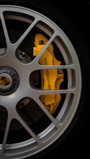 A close-up of a car wheel featuring a silver alloy rim with a yellow brake caliper that has the brand name 'Porsche' written on it. The caliper is highly visible through the spokes of the rim. The central hub displays the Porsche logo.