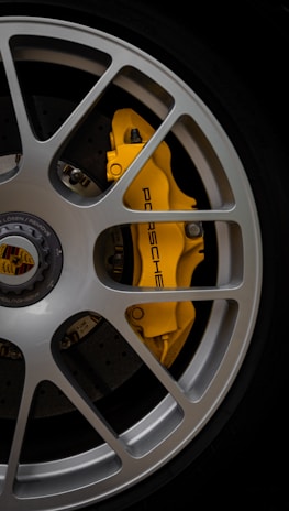 A close-up of a car wheel featuring a silver alloy rim with a yellow brake caliper that has the brand name 'Porsche' written on it. The caliper is highly visible through the spokes of the rim. The central hub displays the Porsche logo.
