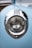 A close-up view of a vintage car's headlight featuring intricate chrome detailing and a soft blue paint finish. The design includes classic pinstriping around the light, and the reflection of trees and sky can be seen on the chrome surface.